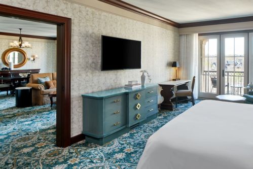 A stylish hotel room featuring a bed, TV, desk, and a view of a balcony with elegant decor and patterned carpet.