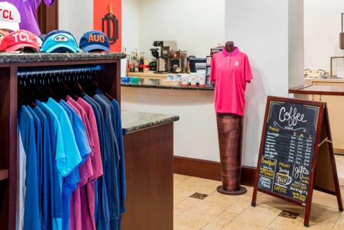 A retail space featuring colorful shirts, a pink polo on a mannequin, and a coffee menu on a chalkboard.