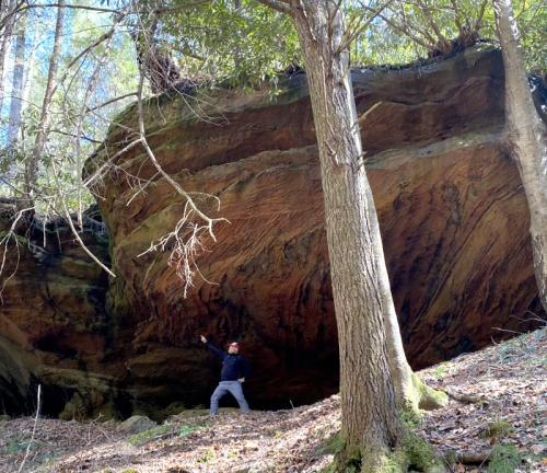 A person stands beside a large rock formation in a forest, surrounded by trees and sunlight filtering through the leaves.