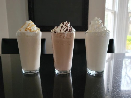 Three tall glasses of milkshakes topped with whipped cream and drizzles, arranged on a dark countertop.