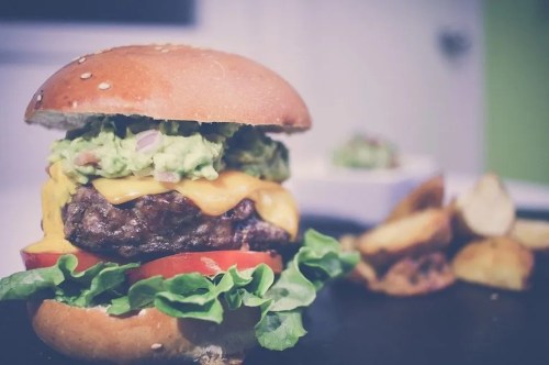 A juicy cheeseburger with lettuce, tomato, and guacamole, served with crispy potato wedges on the side.