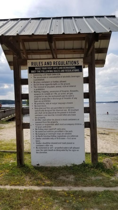Sign listing rules and regulations for a beach area, with a wooden shelter and water in the background.