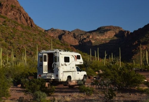 A camper truck parked in a desert landscape with mountains and tall cacti under a clear blue sky.