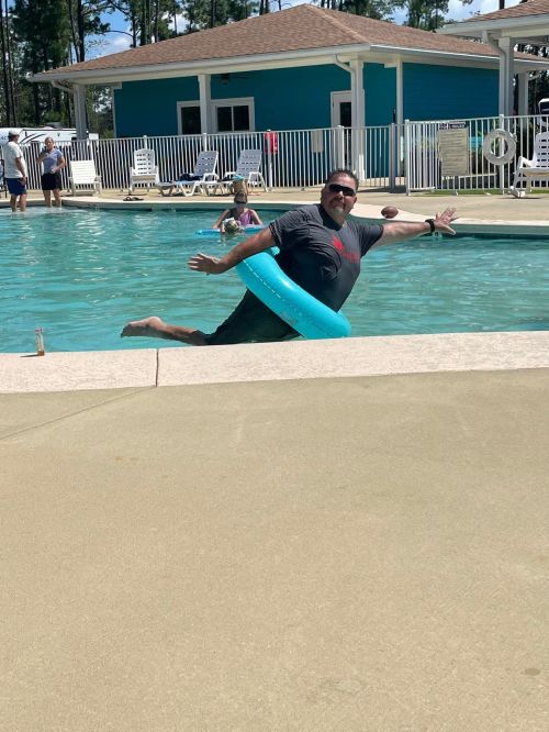 A man in a black shirt playfully leans back in a pool with a blue float, enjoying a sunny day.