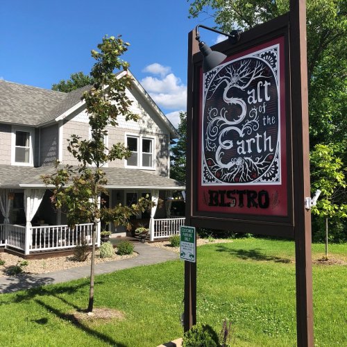 A charming bistro sign in front of a cozy house, surrounded by greenery and blue skies.