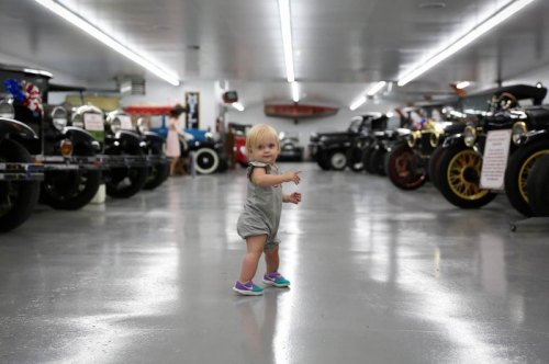 A toddler in a gray outfit stands in a vintage car museum, pointing and surrounded by classic cars.