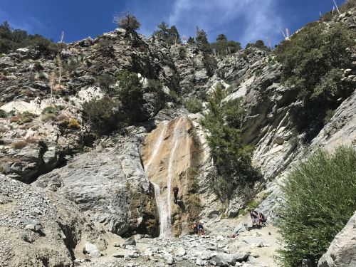 A waterfall cascades down rocky cliffs, surrounded by trees and hikers enjoying the scenic landscape.