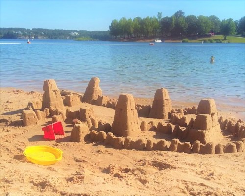 A detailed sandcastle with towers sits on a sandy beach by a calm lake, with a red bucket and yellow shovel nearby.