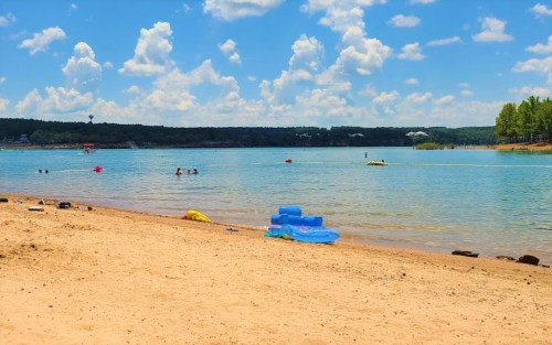 A sandy beach by a calm lake, with people swimming and colorful floats on the water under a blue sky with fluffy clouds.