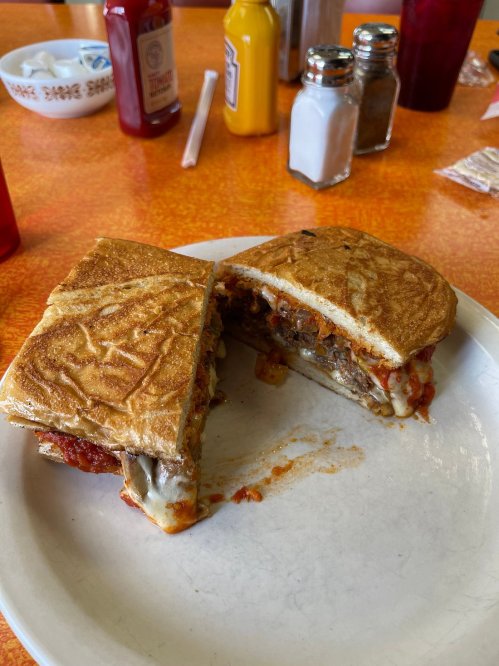 A half-eaten sandwich with melted cheese, meat, and sauce on a plate, surrounded by condiments in a diner setting.