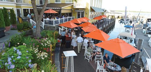 Outdoor dining area with orange umbrellas, flowers, and boats docked nearby, bustling with people enjoying the waterfront.