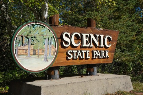 Wooden sign for Scenic State Park featuring a circular illustration of trees and cabins. Surrounded by greenery.