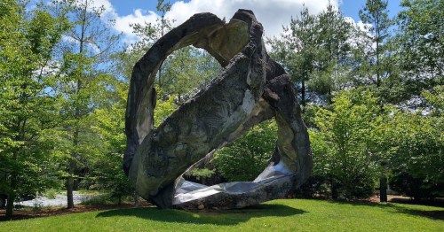 A large abstract sculpture with intertwined forms, surrounded by lush green trees and a blue sky.