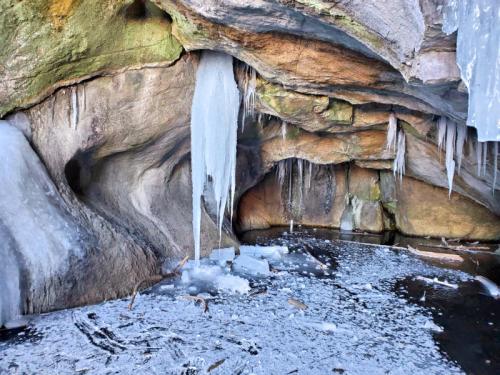 Icicles hang from rocky cave walls above a frozen, icy surface with scattered ice chunks.