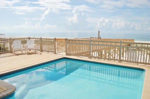 A serene poolside view with wooden deck overlooking the beach and ocean under a bright, cloudy sky.