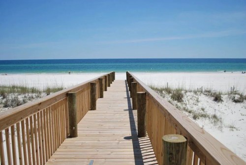 A wooden boardwalk leads to a serene beach with white sand and turquoise waters under a clear blue sky.