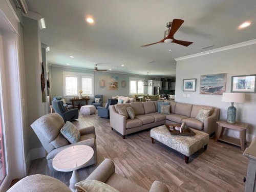 A spacious, modern living room with light-colored furniture, a ceiling fan, and decorative coastal artwork.