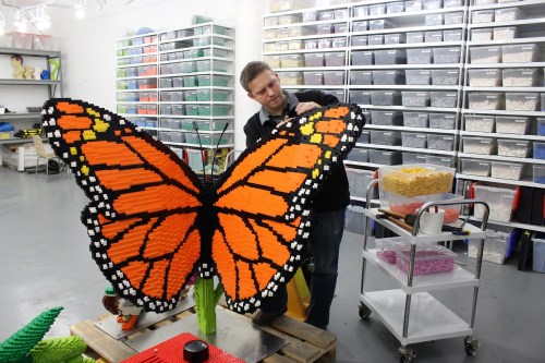 A person assembling a large butterfly sculpture made of colorful LEGO bricks in a workshop filled with storage bins.
