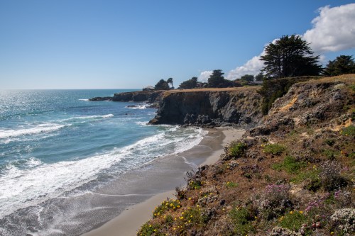 Coastal view with waves crashing on a sandy beach, cliffs, and greenery under a clear blue sky.