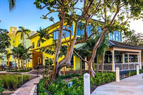 A vibrant yellow building surrounded by lush greenery and palm trees, with a pathway leading to the entrance.