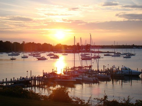 Sunset over a marina with sailboats and reflections on the water, surrounded by trees and a peaceful sky.