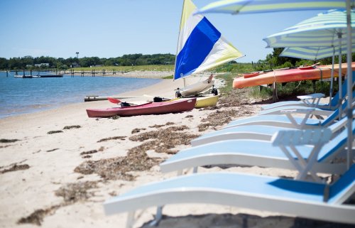 Colorful kayaks and a sailboat on a sandy beach, with lounge chairs and umbrellas in the foreground.
