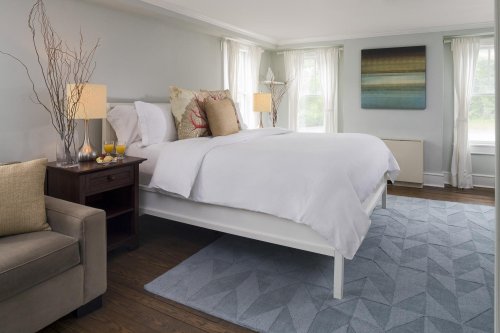 Bright, modern bedroom with a white bed, decorative pillows, a cozy chair, and large windows letting in natural light.
