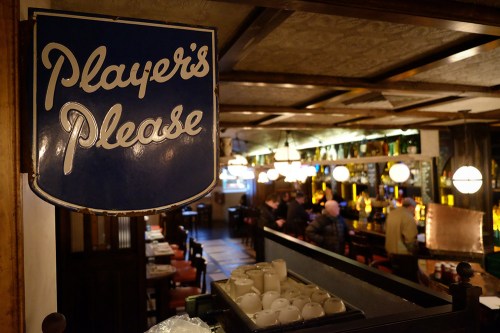 A vintage sign reading "Player's Please" hangs in a cozy bar with patrons enjoying drinks in the background.