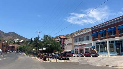 A sunny street scene with buildings, parked cars, and mountains in the background under a clear blue sky.