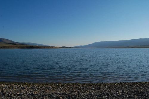 A serene lake surrounded by mountains under a clear blue sky, with a rocky shoreline in the foreground.