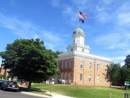 Historic building with a green dome and American flag, surrounded by trees and parked cars under a blue sky.