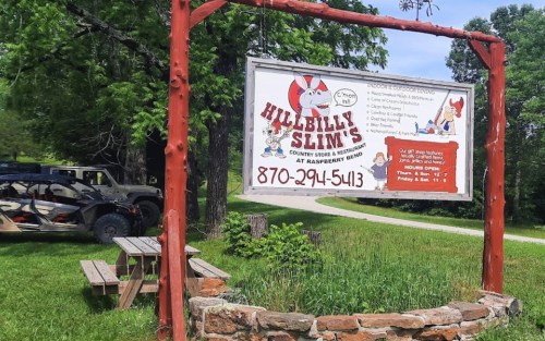 Sign for Hillbilly Slim's restaurant, featuring cartoon graphics, surrounded by greenery and picnic tables.