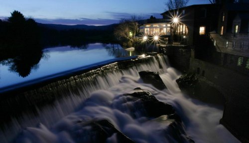 A serene night scene of a waterfall flowing beside a building, illuminated by lights, with a calm river reflecting the sky.