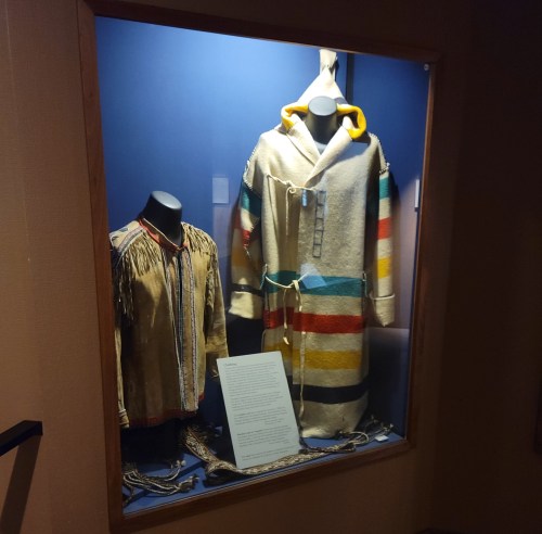 Display case featuring traditional garments, including a fringed shirt and a colorful striped hooded coat.