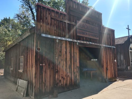 A rustic wooden blacksmith shop with a sign reading "Harmon & Harrison," surrounded by trees and sunlight.
