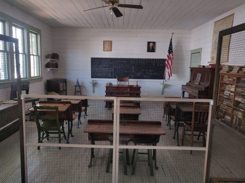 A vintage classroom with wooden desks, a chalkboard, a piano, and a U.S. flag, enclosed by a wire mesh barrier.