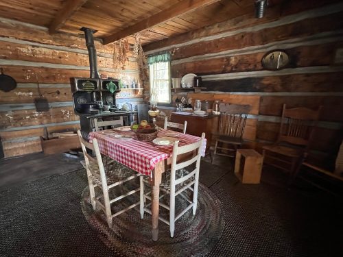 A rustic kitchen with a wooden table set for a meal, surrounded by chairs and a vintage stove in a log cabin.