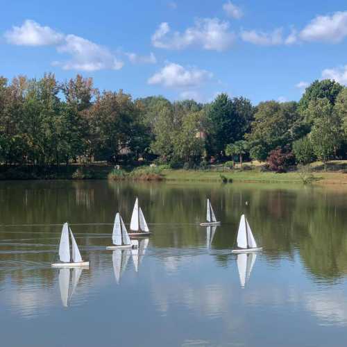 Model sailboats glide across a calm lake, reflecting trees and a blue sky with fluffy clouds.