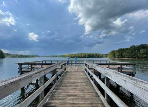 Two people stand on a wooden pier overlooking a calm lake under a cloudy sky.