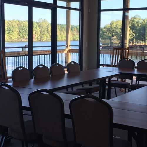 A meeting room with several tables and chairs, overlooking a lake and trees through large windows.