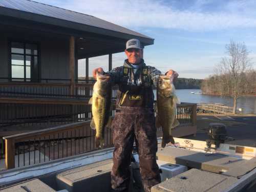 A person in fishing gear holds two large bass fish, standing on a boat with a lake and building in the background.