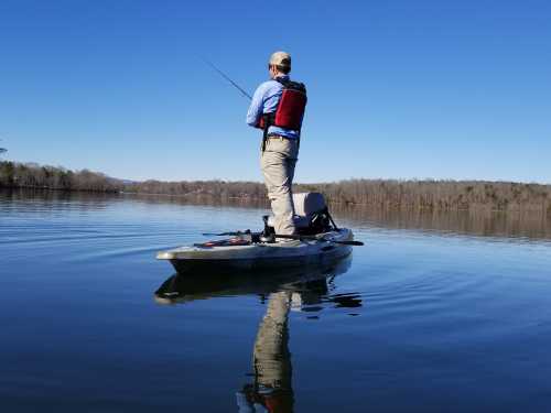 A person fishing from a kayak on a calm lake, surrounded by trees under a clear blue sky.