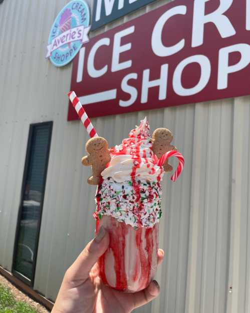 A festive milkshake topped with whipped cream, sprinkles, and gingerbread cookies, held in front of an ice cream shop sign.