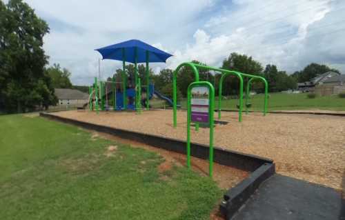 A colorful playground with green equipment and a blue shade structure, set in a grassy area under a cloudy sky.