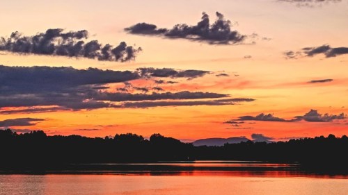 A serene sunset over a calm lake, with vibrant orange and purple hues reflecting on the water and clouds in the sky.