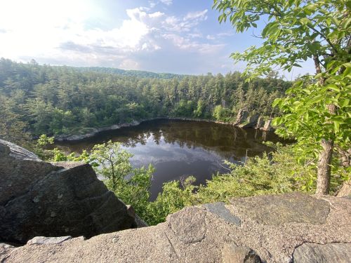 A serene view of a calm lake surrounded by lush greenery and rocky cliffs under a partly cloudy sky.