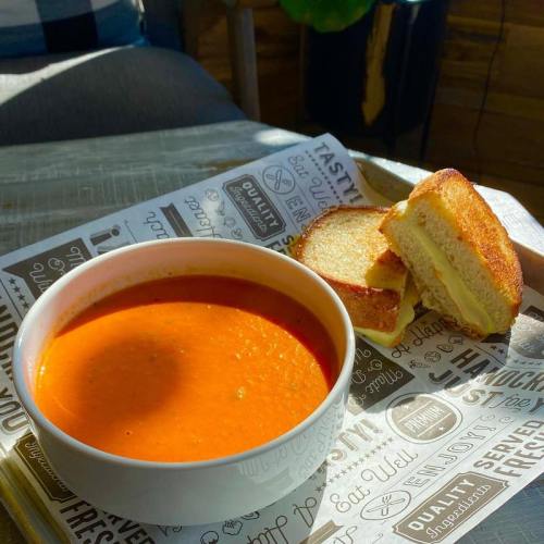 A bowl of tomato soup next to a plate with two grilled cheese sandwiches on a patterned table.