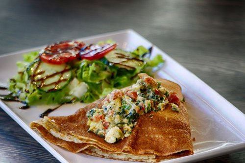 A plate featuring a stuffed crepe with vegetables and a side salad of lettuce and tomatoes drizzled with balsamic.