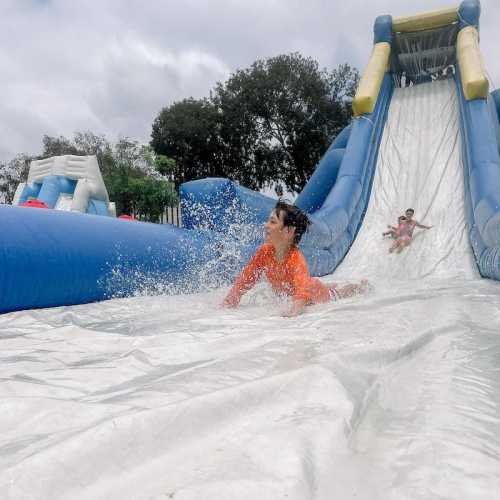 A child in an orange swimsuit slides down a water slide, splashing into a pool, with another child sliding behind.