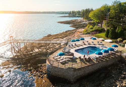 Aerial view of a pool by the ocean, surrounded by lounge chairs and umbrellas, with a rocky shoreline and sunset.